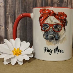 Peut inclure: Mug en c&eacute;ramique blanche avec une anse et un bord rouges, repr&eacute;sentant un carlin portant des lunettes rouges et un bandana &agrave; pois. Le mug porte l'inscription "Pug Mom" et est &agrave; c&ocirc;t&eacute; d'une marguerite en papier blanche.