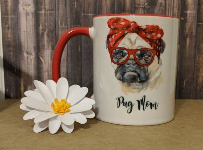 Peut inclure: Mug en c&eacute;ramique blanche avec une anse et un bord rouges, repr&eacute;sentant un carlin portant des lunettes rouges et un bandana &agrave; pois. Le mug porte l'inscription "Pug Mom" et est &agrave; c&ocirc;t&eacute; d'une marguerite en papier blanche.