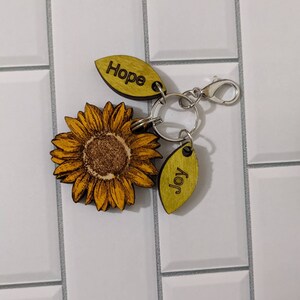 May include: A wooden sunflower keychain with a silver key ring and two wooden tags that say "Hope" and "Joy".