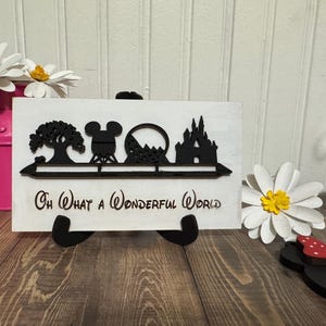 Può includere: Un cartello di legno bianco con sagome nere di un albero, la testa di Mickey Mouse, montagne russe e un castello. Il cartello recita "Oh What a Wonderful World".