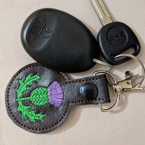 May include: A brown leather keychain with a green and purple embroidered thistle design. The keychain is attached to a silver key ring and a black car key fob.
