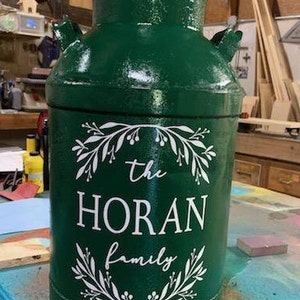 Puede incluir: Un cántaro de leche verde oscuro con un diseño floral blanco y el texto "the HORAN family". El cántaro tiene una parte superior redondeada y dos asas laterales. Está sobre una superficie azul y marrón.