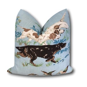 May include: A light blue pillow with a pattern of brown and white dogs running through a field of green grass and blue flowers. There are also small blue birds flying in the field.