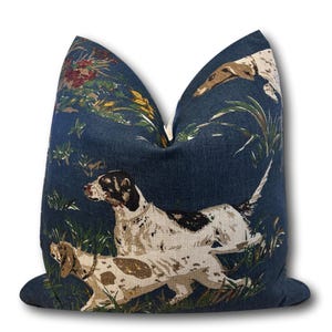 May include: A blue throw pillow with a hunting dog pattern. The dogs are running through tall grass and are white with brown spots. The background is a dark blue.