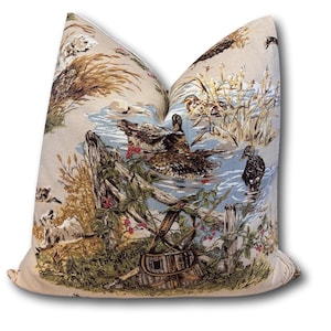 May include: Decorative throw pillow with a nature-themed print. The pillow features a detailed illustration of ducks in a pond, with a wooden fence, reeds, and a woven basket. The color palette includes beige, blue, brown, and green.
