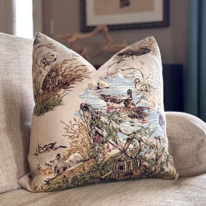 May include: Beige decorative pillow with a pastoral scene featuring ducks, rabbits, and a pond. The pillow has a detailed print with brown, green, and blue accents. The pillow is placed on a beige sofa.