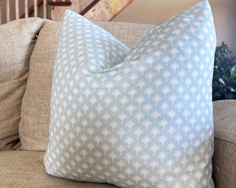May include: A decorative throw pillow with a light blue background and a white, repeating floral pattern. The pillow is placed on a beige sofa, with a wooden staircase in the background. The pillow adds a touch of color and pattern to the neutral-toned living space.