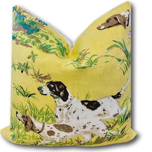 May include: Yellow pillow with a white border featuring a repeating pattern of brown and white dogs running through tall grass. The dogs are in a hunting scene with green foliage and brown branches.