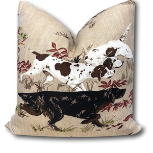 May include: A beige pillow with a brown and white dog and a black dog running through a field of red and green foliage.