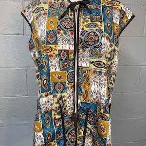 May include: A sleeveless vest with a colorful patchwork design. The vest features a zipper closure and a black trim. The pattern includes paisley and floral motifs in shades of blue, yellow, red, and white. A black bow is at the neckline.