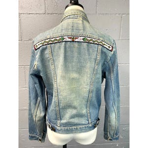 May include: A light blue denim jacket with a beaded design on the back. The design features a snake and a bird.