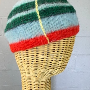 May include: A colorful, hand-knitted hat with horizontal stripes of green, blue, and red. The hat has a yellow pom-pom on top and is displayed on a wicker mannequin head. The hat's brim is red.