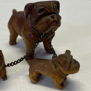 May include: Three wooden bulldog figurines, one larger than the other two, connected by a metal chain. The larger bulldog is brown with black markings, while the smaller bulldogs are a lighter brown.