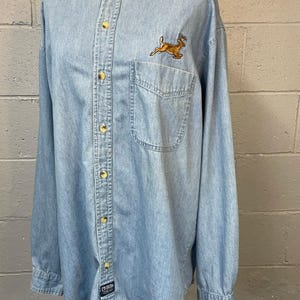 May include: Pale blue denim shirt with a mandarin collar and long sleeves. It has a small chest pocket and a running deer embroidered on the upper left. The shirt has gold-coloured buttons and a small label near the bottom.