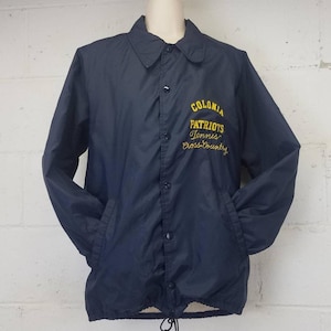 May include: Navy blue nylon jacket with a button-up front, collar, and pockets. The jacket features the embroidered text "COLONIA PATRIOTS Tennis Cross-Country" in yellow script. A drawstring is visible at the bottom hem.