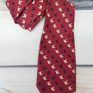May include: A burgundy tie with a repeating pattern of white and black bird silhouettes. The tie is displayed on a light-colored wooden surface, showcasing its design and color contrast. The tie is a classic accessory.