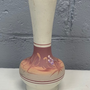 May include: A small, white ceramic vase with a brown and orange painted design. The vase has a narrow neck and a wide base. The design features stylized flowers and leaves.