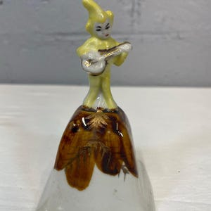 May include: A vintage ceramic bell featuring a musician figure. The figure is dressed in yellow and plays a small guitar. The bell has a white base with brown and gold floral accents, creating a whimsical and decorative piece.