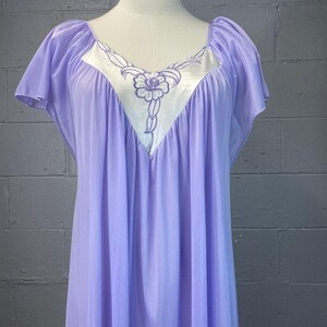 May include: A light purple nightgown featuring short flutter sleeves and a V-neckline. The bodice includes a white triangular inset panel decorated with purple floral embroidery. The garment is made of a sheer, flowing fabric with gathered details.