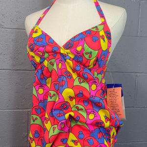 May include: A halter-neck swimsuit with a vibrant, psychedelic floral pattern in shades of pink, yellow, green, and blue. The swimsuit is displayed on a mannequin against a gray brick wall. A tag is attached to the garment.