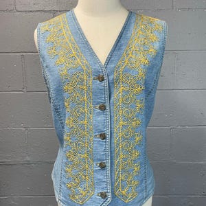 Peut inclure: Un gilet en jean bleu clair avec des motifs floraux brodés jaunes. Le gilet a un col en V et un devant boutonné.