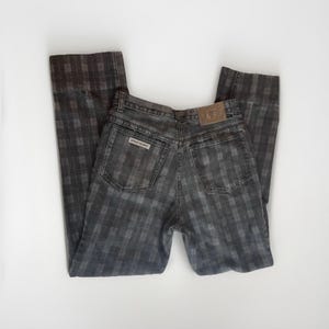 May include: Vintage gray plaid jeans with a straight leg. The jeans feature a classic five-pocket design, a leather patch, and the brand name "Sergio Valente" on the back pocket. The plaid pattern is a mix of gray and black.