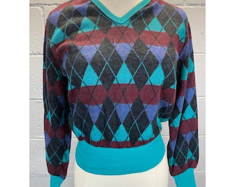 May include: A long-sleeved sweater with a v-neck and a diamond pattern in shades of teal, purple, black, and burgundy. The sweater has a turquoise band at the bottom.