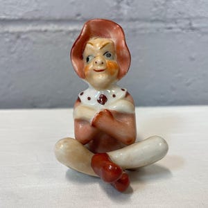May include: A vintage ceramic figurine of a seated elf with a mischievous expression. The elf wears a pink hat and outfit with a white collar and red accents. The figurine is hand-painted with detailed features.