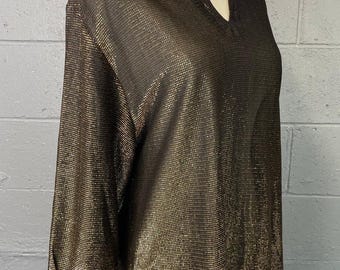 May include: A long-sleeved, metallic gold and brown blouse with a V-neck collar. The fabric has a textured, grid-like pattern. The blouse is displayed on a mannequin against a gray brick wall.