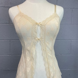 Könnte beinhalten: Elfenbeinfarbenes Spitzen-Camisole mit zarten Blumenmustern und gewellten Kanten. Verfügt über dünne Träger und kleine Schleifenakzente an der Brust und in der Mitte. Das Camisole wird auf einer Schaufensterpuppe präsentiert.