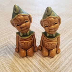 May include: Two ceramic figurines of elves with green hats and brown bodies. They are sitting with their legs crossed and have a mischievous expression on their faces.
