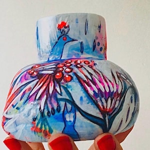 May include: A hand-painted ceramic vase with a wide base and a cylindrical neck. The vase features a vibrant peacock design with blue, red, and purple hues. The artwork includes a peacock, flowers, and abstract shapes, creating a unique decorative piece.
