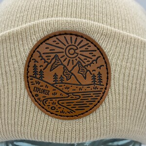 May include: A beige knitted beanie with a brown leather patch featuring a mountain scene, a sun with a 'C' inside, and the word 'EXPLORER'.