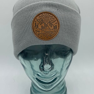 May include: Gray knitted beanie with a brown leather patch featuring a mountain scene, a sun, and the word "Explorer".