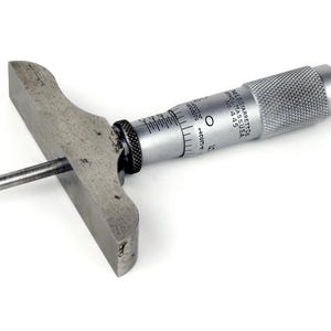 May include: A depth micrometer tool with a silver body and a dark gray handle. The tool has a flat base and a long, thin rod extending from it. The micrometer has measurement markings and is used for precise depth measurements.