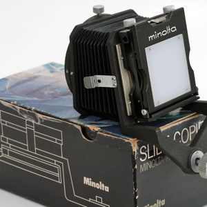 May include: A black Minolta Slide-Copier with a bellows and a white screen. The copier is sitting on a black box with the Minolta logo.