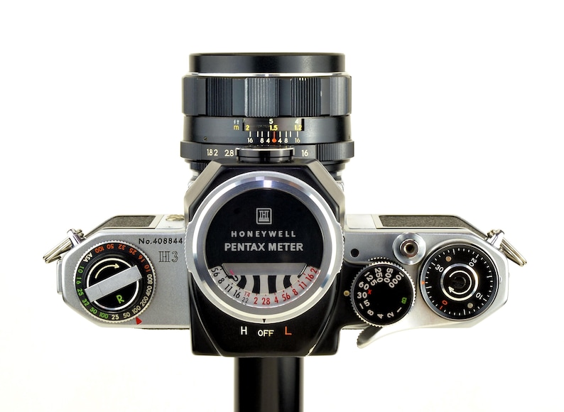 Może przedstawiać: Zabytkowy aparat fotograficzny Honeywell Pentax z czarnym obiektywem i srebrnym korpusem. Aparat posiada r&oacute;żne pokrętła, miernik i napis "Honeywell Pentax Meter". Obiektyw posiada oznaczenia pomiarowe.