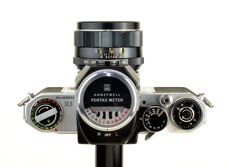 Może przedstawiać: Zabytkowy aparat fotograficzny Honeywell Pentax z czarnym obiektywem i srebrnym korpusem. Aparat posiada r&oacute;żne pokrętła, w tym pokrętło ASA i miernik Pentax. Obiektyw posiada oznaczenia pomiarowe. Aparat stoi na białym tle.