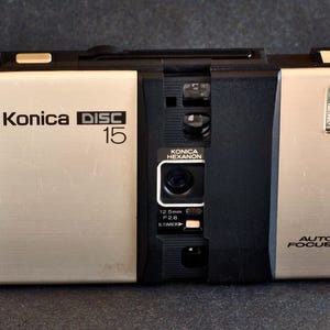 May include: A silver and black Konica Disc 15 camera featuring a central black panel with a Konica Hexanon 12.5mm F2.8 lens, an integrated flash unit on the right, and Auto Focus text. The camera body has a rectangular shape with a metallic finish.