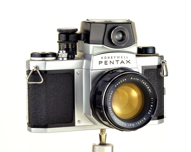 Może przedstawiać: Zabytkowy aparat fotograficzny Honeywell Pentax ze srebrnym korpusem, czarnymi akcentami i wyraźnym obiektywem. Obiektyw oznaczony napisem "Auto-Takumar 1:1.8/55". Aparat posiada wizjer, pokrętła i teksturowany uchwyt. Widoczny jest napis "Honeywell Pentax".
