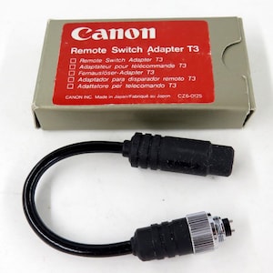 May include: A Canon Remote Switch Adapter T3 in its original gray cardboard box with a red label. The adapter features a short black cable with a black plastic connector on one end and a black plastic connector with a silver metal ring on the other.