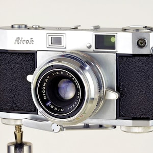 May include: A vintage Ricoh camera with a silver body and black textured grips. The lens is labeled "RICOMAT 1:2.8" and "RIKEN". The camera has a classic design, with a viewfinder and various dials.