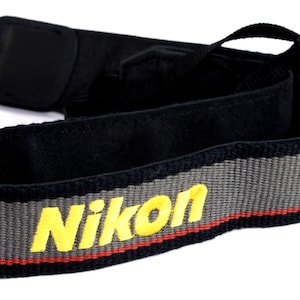 Può includere: Cinghia per fotocamera nera con una striscia grigia e rossa e la parola "Nikon" in lettere gialle.
