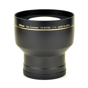 Könnte beinhalten: Schwarzer Nikon Tele Converter TC-E17ED 1.7x Objektiv. Das Objektiv hat ein goldenes Band mit dem Nikon-Logo und Text. Hergestellt in Japan. Geriffelter Sockel und breite Öffnung.