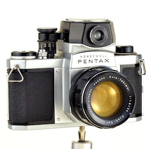 Może przedstawiać: Zabytkowy aparat fotograficzny Honeywell Pentax ze srebrnym korpusem, czarnymi akcentami i wyraźnym obiektywem. Obiektyw oznaczony napisem "Auto-Takumar 1:1.8/55". Aparat posiada wizjer, pokrętła i teksturowany uchwyt. Widoczny jest napis "Honeywell Pentax".