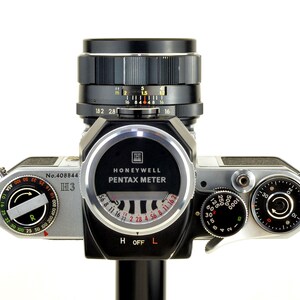 Może przedstawiać: Zabytkowy aparat fotograficzny Honeywell Pentax z czarnym obiektywem i srebrnym korpusem. Aparat posiada r&oacute;żne pokrętła, miernik i napis "Honeywell Pentax Meter". Obiektyw posiada oznaczenia pomiarowe.