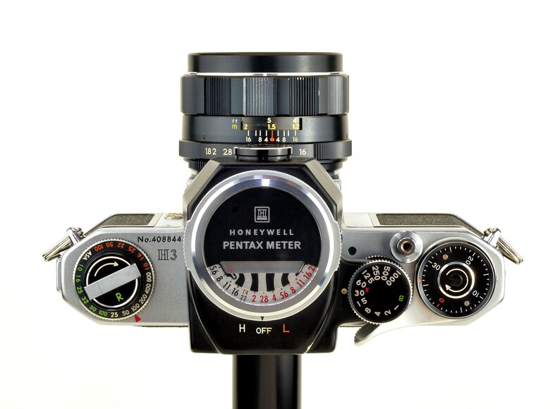 Może przedstawiać: Zabytkowy aparat fotograficzny Honeywell Pentax z czarnym obiektywem i srebrnym korpusem. Aparat posiada r&oacute;żne pokrętła, miernik i napis "Honeywell Pentax Meter". Obiektyw posiada oznaczenia pomiarowe.