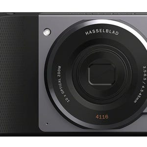 Op de afbeelding: Een zwarte camera-accessoire met een getextureerde grip, een grote ronde lens en een oranje ontspanknop. Op de lens staat de tekst HASSELBLAD, 10x OPTICAL ZOOM, 3.5-6.5 / 4.5-45mm en 4116. Een zilverkleurig H-logo is rechtsonder zichtbaar.
