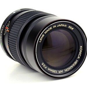 May include: A black Konica Hexanon AR 135mm f3.5 lens with gold lettering. The lens is made in Japan and has a 55mm filter size.