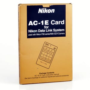 Op de afbeelding: Een gouden Nikon AC-1E Card doos voor het Nikon Data Link System. De doos heeft een donkerblauw rechthoek met het Nikon-logo en productinformatie. Een diagram van de kaart en zijn functies is ook zichtbaar.